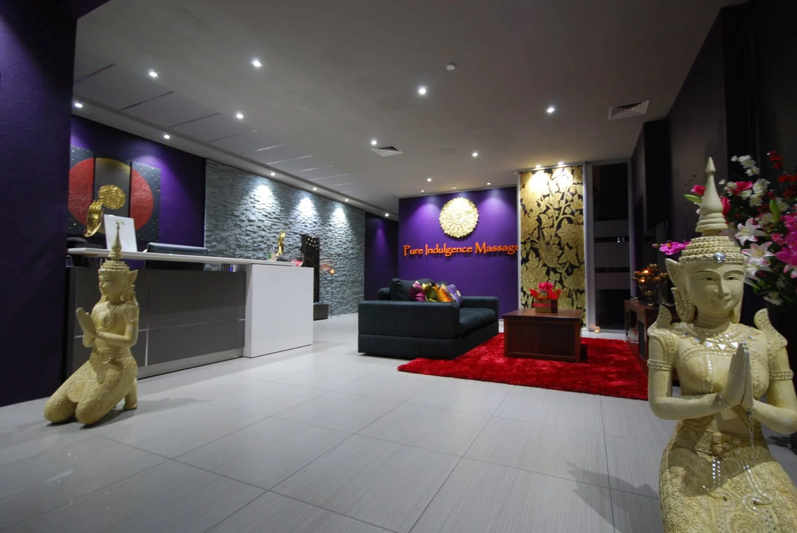 Reception — gold Thai deva statues, glowing Pure Indulgence Massage wordmark on deep purple wall