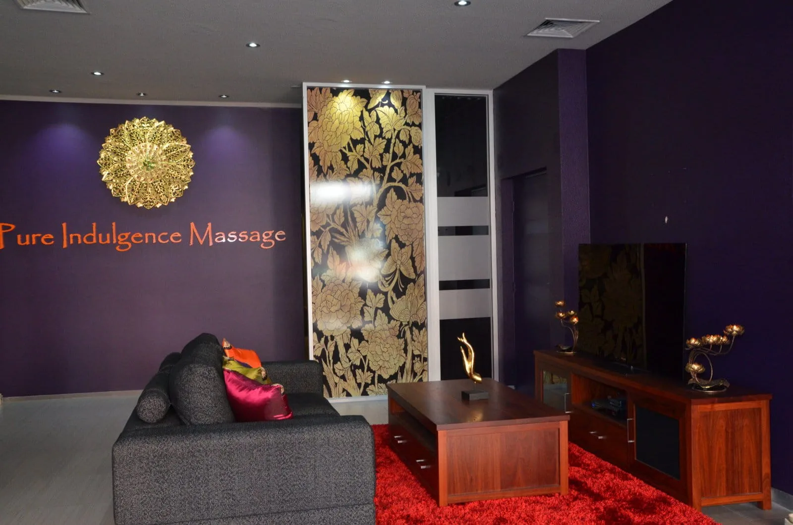 Waiting lounge — gold floral wall panel, grey sofa on red shag rug, candlelight and lotus ornaments